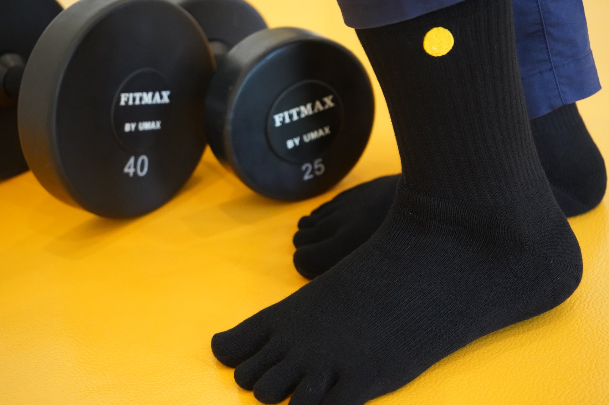Black Fitmax dumbbells with a person wearing black toe socks on a yellow background