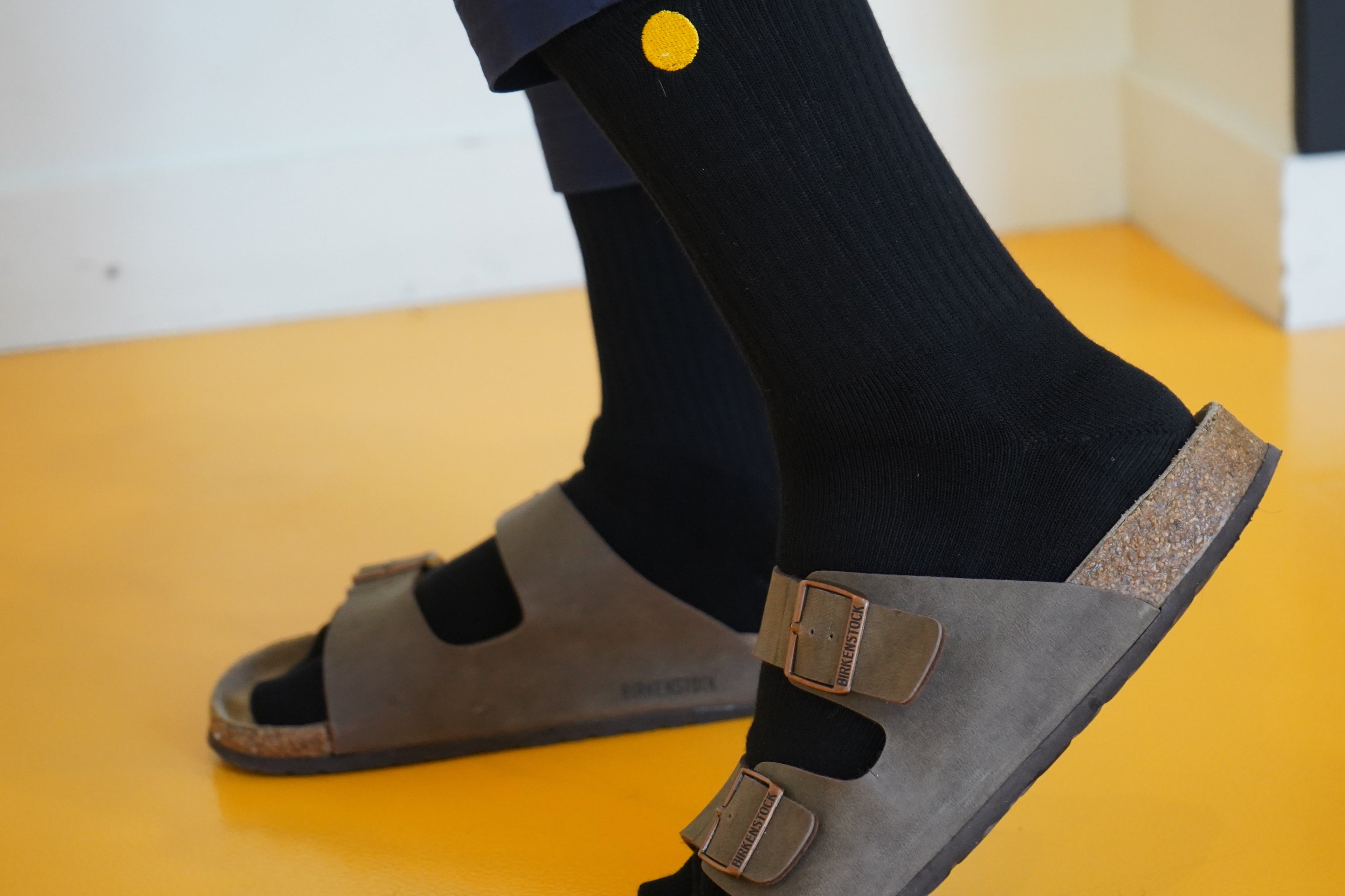 Person wearing brown sandals with valuura Toe Socks on a yellow floor.